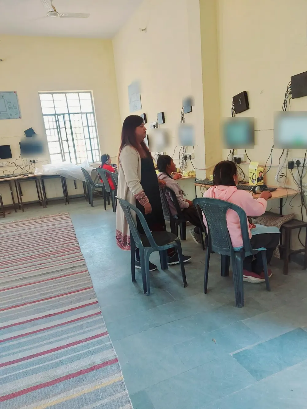 A teacher stands beside students working at computers in a school lab.